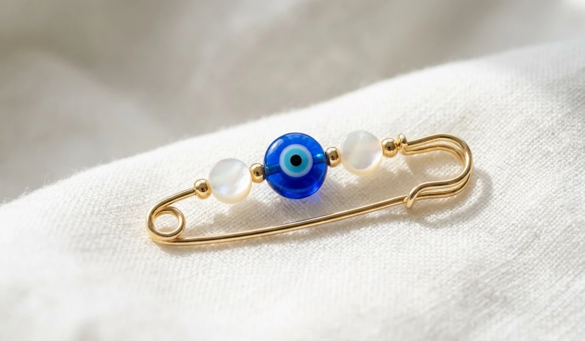 The Spiritual Meaning and Protection of Evil Eye Baby Pins - Buddha Bodhis A high-quality close-up of a gold baby safety pin with a blue glass evil eye charm on white linen.